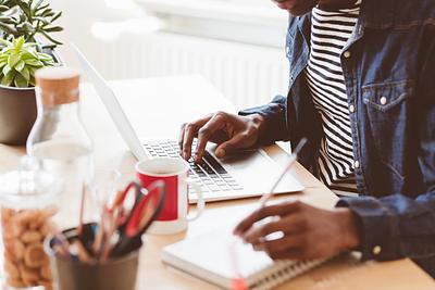 Man sitting at a desk, working on an open laptop wile also writing in a paper notebook. His head is out of frame. 