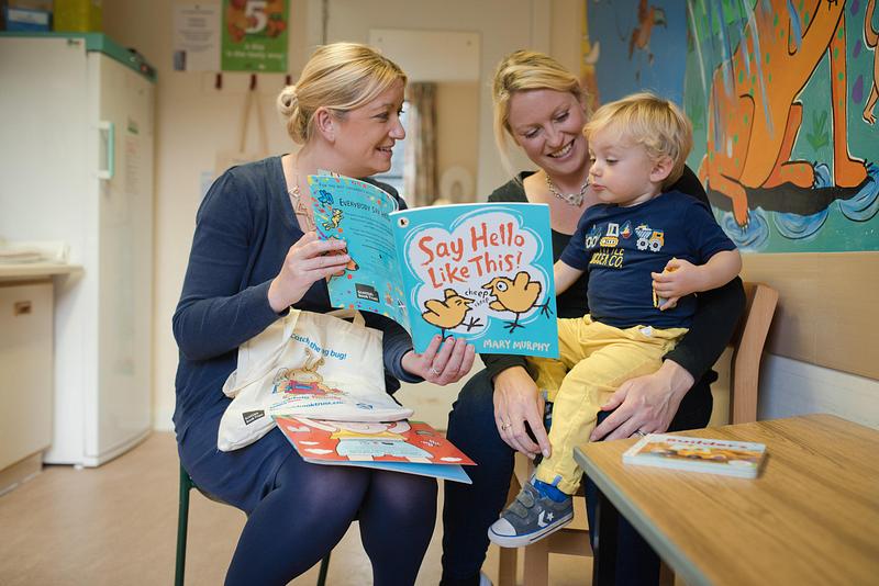 Two women reading 'Say Hello Like This' from a Bookbug Bag to a young boy 