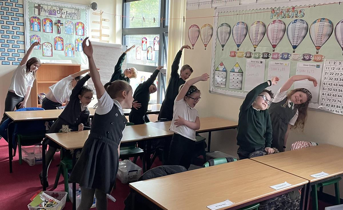 Group of primary school children standing behind desks in  a classroom, all raising one arm and bending to one side in a balletic pose.