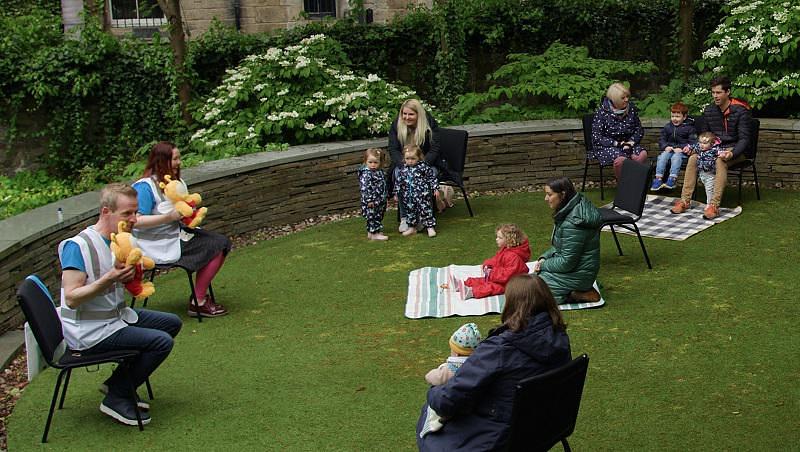 Four families with young children seated outside in a garden watching a man and a woman holding Bookbug dolls