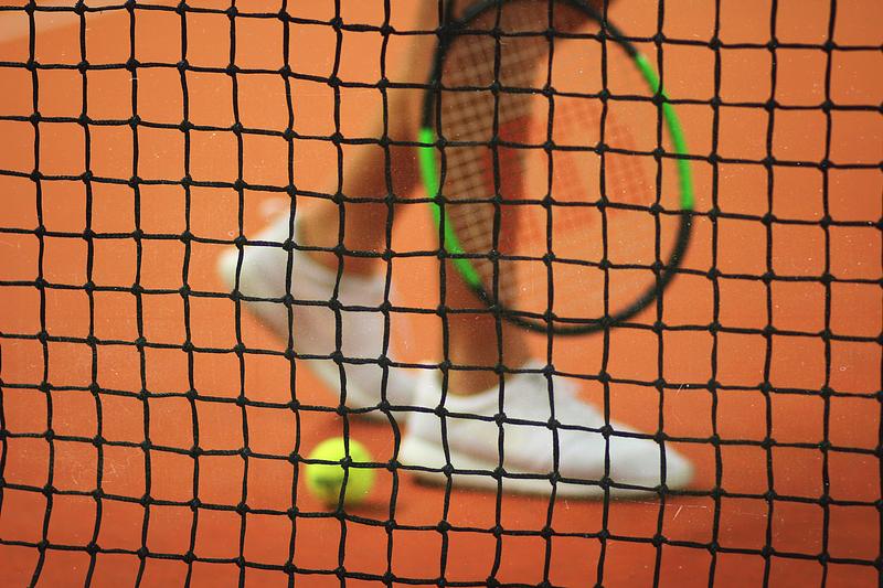 View through a tennis net of a person's legs, racket and ball on the court.