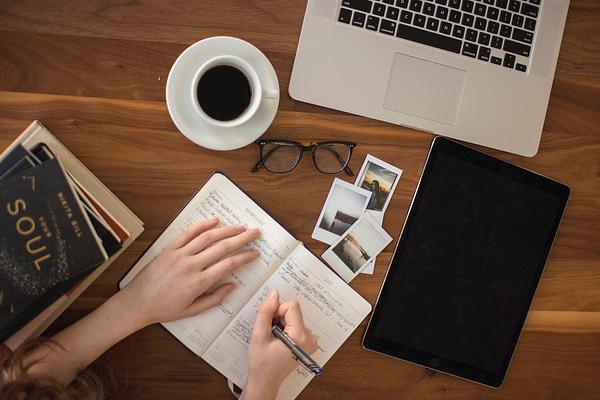Close up of someone writing in a notebook at a wooden desk, covered with books, a laptop, photos and glasses. Close up of someone writing in a notebook at a wooden desk, covered with books, a laptop, photos and glasses.