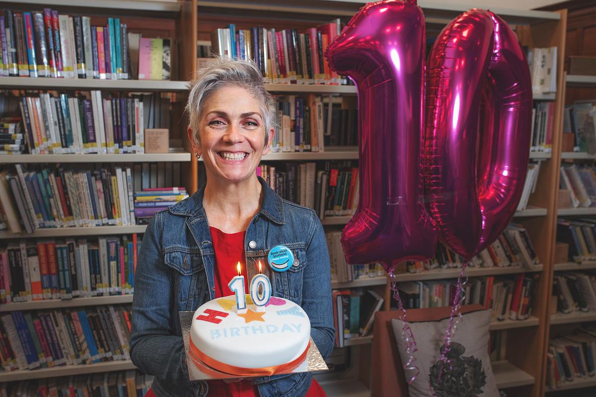 Denise Mina holding birthday cake next to 10 balloons and bookshelves at Glasgow Women's Library