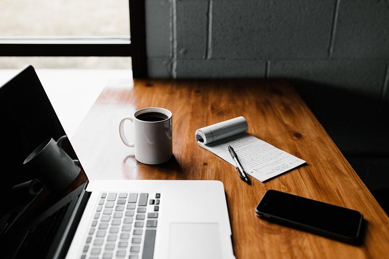 A laptop, mug of coffee, phone and flip notebook on a desk
