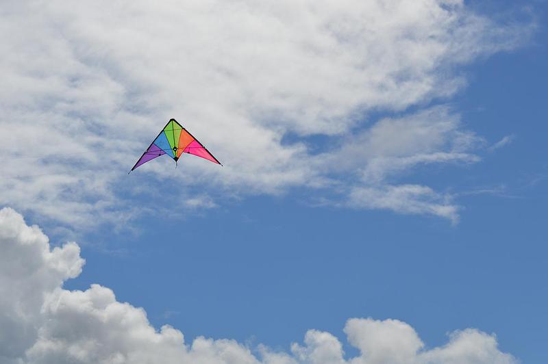 Brightly coloured, triangle shaped kite flying in a blue sky with fluffy white clouds