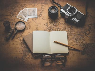 A blank notebook, pencil, camera, spectacles and magnifying glass are arranged on yellowed map of the world