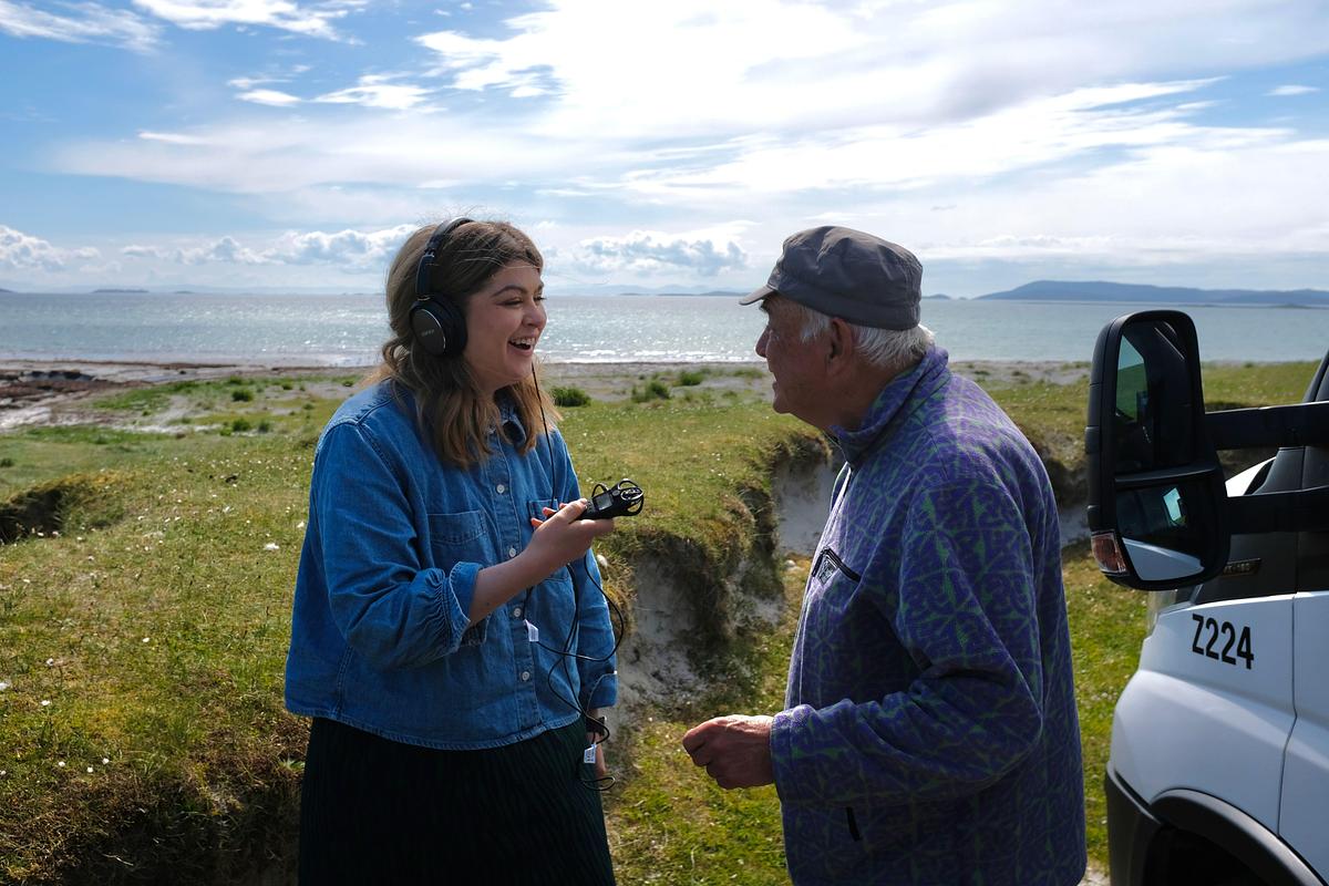A young woman in headphones and holding a microphone, talking to an old man with a beach in the background