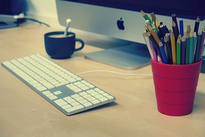 A red pencil pot filled with an assortment of coloured pencils sitting on a desk. There is also a mac computer keybaord and part of a screen visable as well as a cup of coffee in the background. 