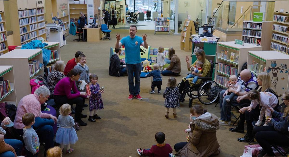 Bookbug Session Leader standing with children their family and  carers in a library