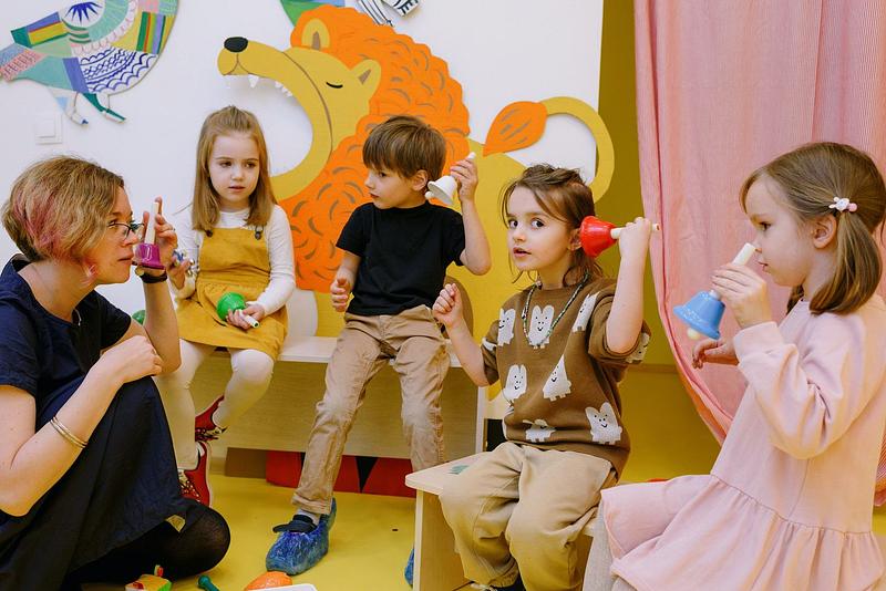 A group of children and a woman holding up colourful bells to their ears to listen