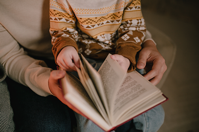 Child in a brown jumper sitting on an adult's lap and sharing a book