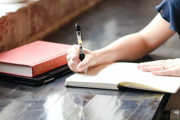 A hand writing in a notebook with another book and tablet nearby on the table