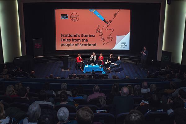 A panel of speakers sitting around a table on a stage. Behind them, the screen reads 'Scotland's Stories: Tales from the people of Scotland'