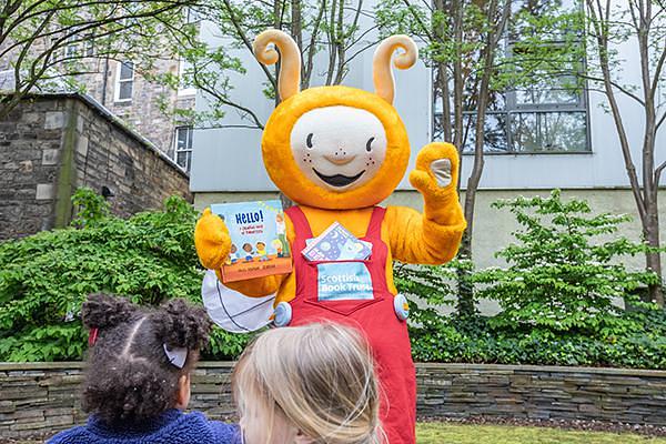 Bookbug in Scottish Book Trust's garden, holding a book called Hello and waving to the camera
