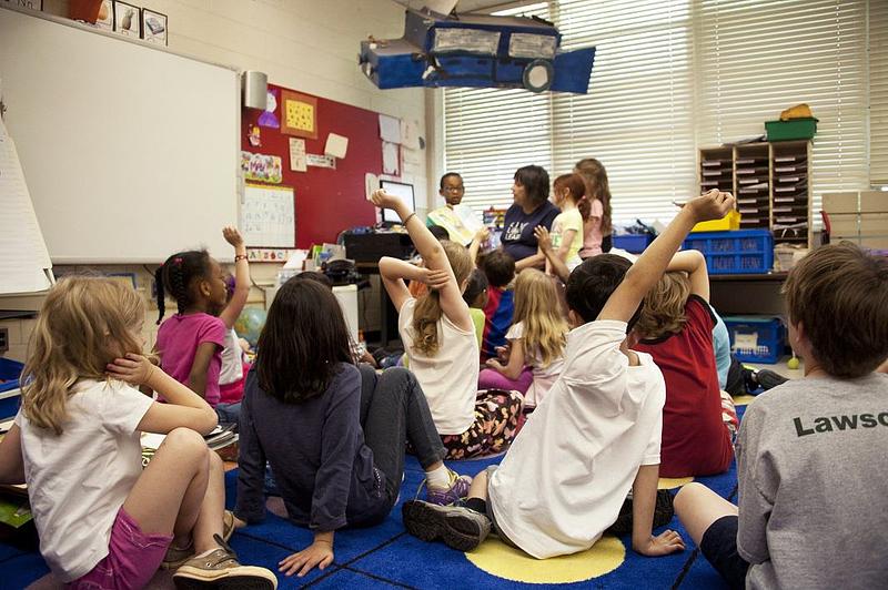 Pupils gathered on the floor facing an author reading a book, some have their hands up