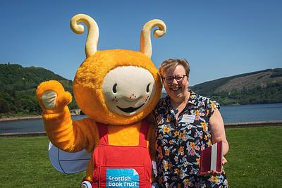 Bookbug waving to the camera with an arm around Janice Middleton, Bookbug Hero Award winner 2023. Janice is holding her trophy and behind them is a loch and hills landscape