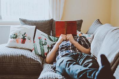 A person relaxing on a corner sofa, their face covered by holding up a book cover