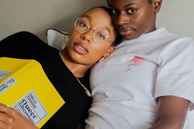 Woman in black shirt with yellow book titled "Queer Intentions" lounging beside woman in white polo shirt