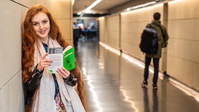 Len Pennie leaning against a wall, smiling at the camera while holding a Book Week Scotland Adventure book. A silver ticket with "25" on it is peeking from the top.