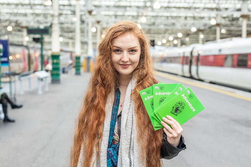 Len Pennie standing in a train station holding four bright green books in a fan. The books are titled 'Adventure' and have a boot print and Book Week Scotland logo on the front.