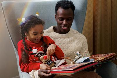 A young girl wearing a Christmas jumper sits on a man's lap reading a pop-up book
