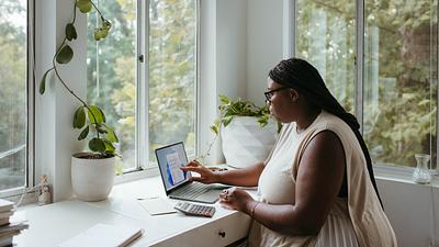 A Black woman with long waist-length braids sits at a table on a laptop