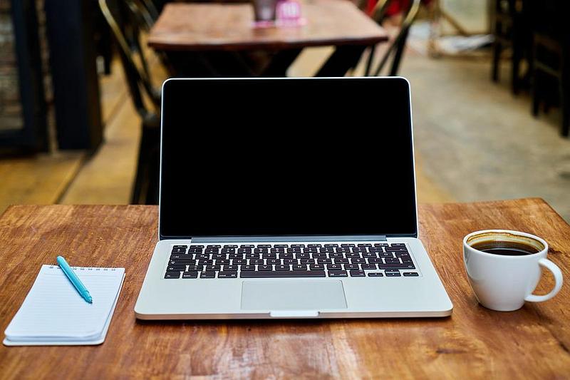 A laptop on a table with  a notepad, pen and a cup of coffee